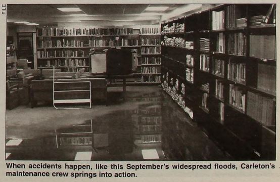 A photograph of flood waters in a room with books, bookshelves, chairs and tables, which have been moved away from the water.