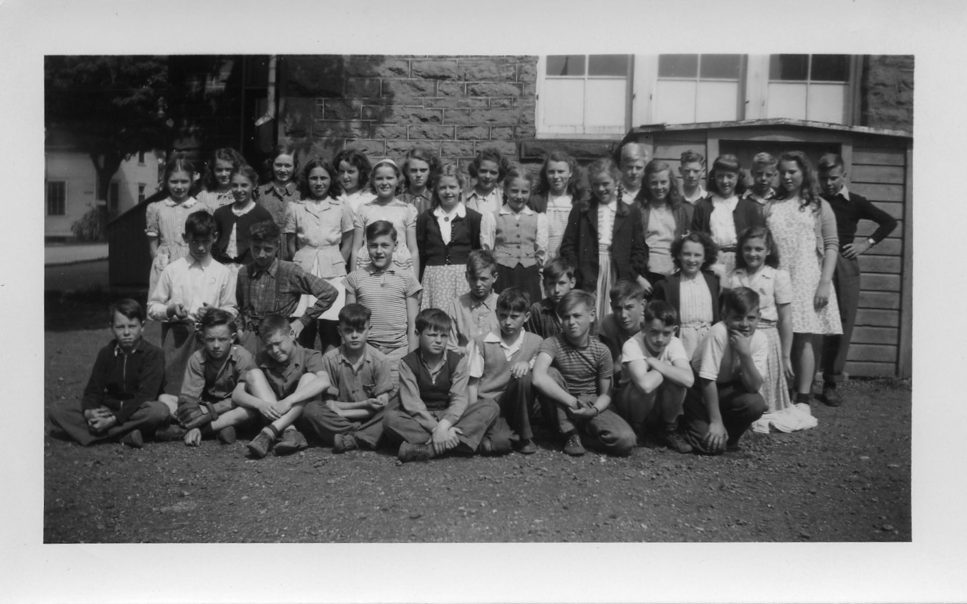 Black and white photograph of a class portrait outside Victoria School. There are four rows. The front row is sitting, the second row is kneeling, and the back rows are standing. It is boys and girls. The photograph is taken at the side of the building. The entrance to the basement bunker is behind them.